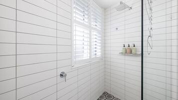 A modern bathroom with white tiled walls, a shower with a glass door, and a window with blinds.