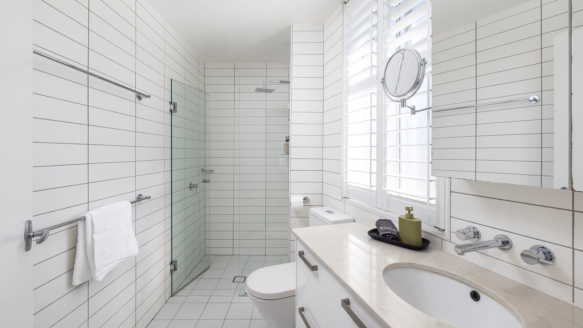 A modern bathroom with white tiled walls, a glass shower enclosure, a white toilet, a sink with a mirror, and a window with blinds.