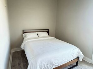 Ein Einzelbett mit Holzrahmen und weißer Bettwäsche in einem Zimmer mit schlichten Wänden und einem Fliesenboden.