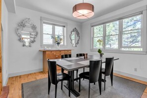 Comedor con mesa, sillas, una lámpara colgante y dos ventanas con vistas a árboles.