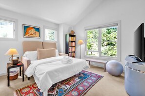 Un dormitorio moderno con cama, mesitas de noche, una estantería, televisor y un amplio ventanal con vistas a árboles.