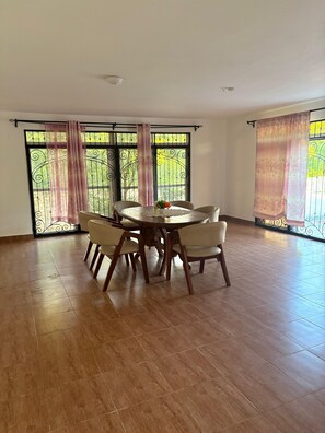 Een ruime eethoek met een ronde tafel en zes stoelen, grote ramen met decoratieve gordijnen, en uitzicht op groen buiten.