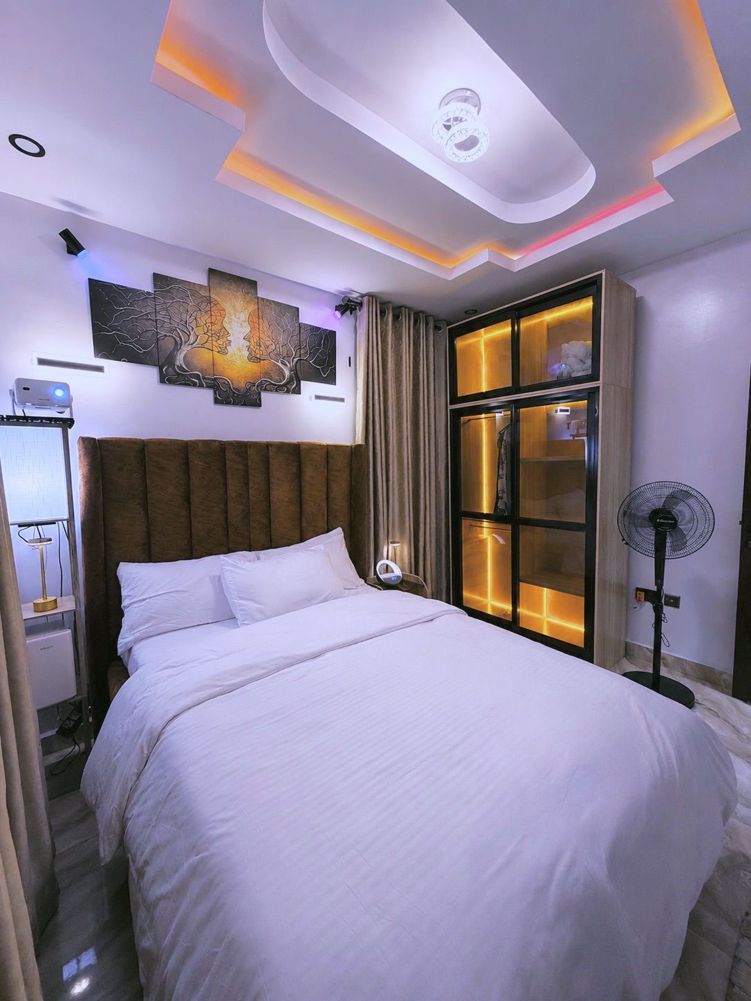 A modern hotel room with a large bed, a ceiling light fixture, a wall-mounted artwork, and a window with curtains.