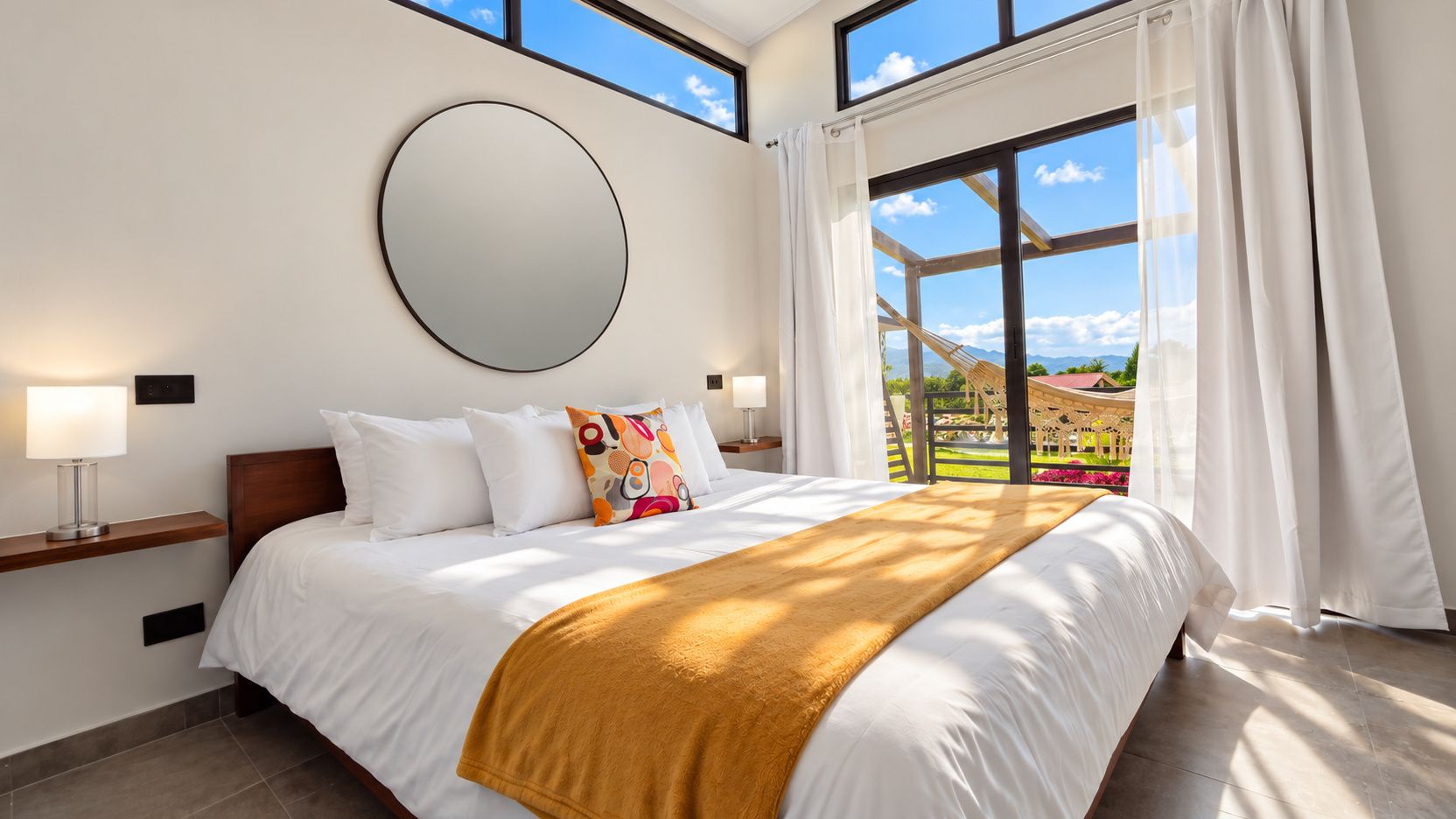 A hotel room with a large bed, a round mirror on the wall, and a view of the outdoors through a large window.