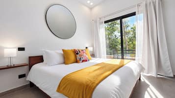 A hotel room with a large bed, a bedside table with a lamp, a round mirror on the wall, and a window with curtains.