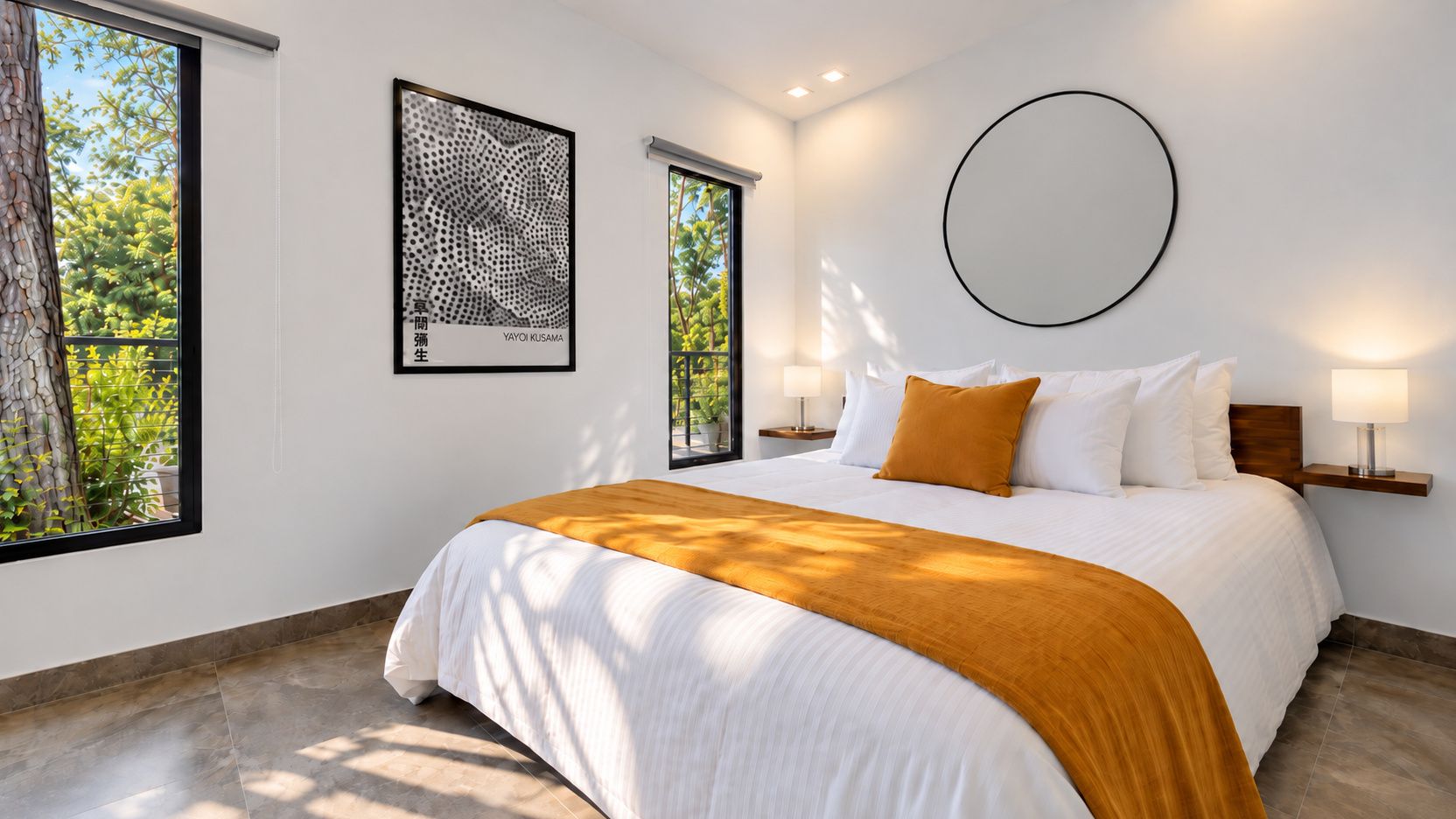 A hotel room with a large bed, two bedside tables with lamps, a round wall decoration, and two windows with a view of greenery.