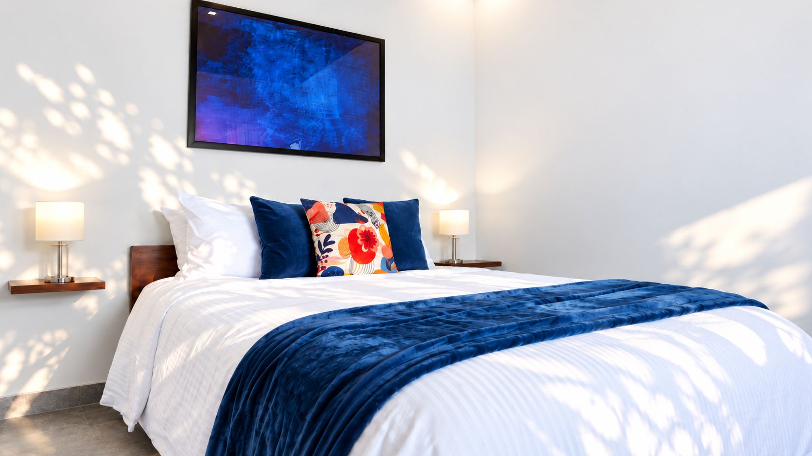 A neatly made bed with a blue blanket, two pillows, and a colorful throw pillow. There are two bedside lamps on wooden nightstands.