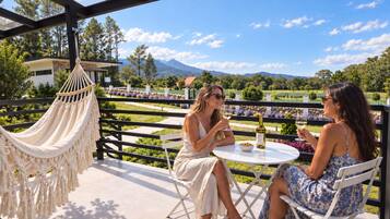 Two people seated at a round table with a bottle of wine and a bowl of nuts, under a white canopy with a macrame hanging chair, overlooking a landscaped garden and distant mountains.