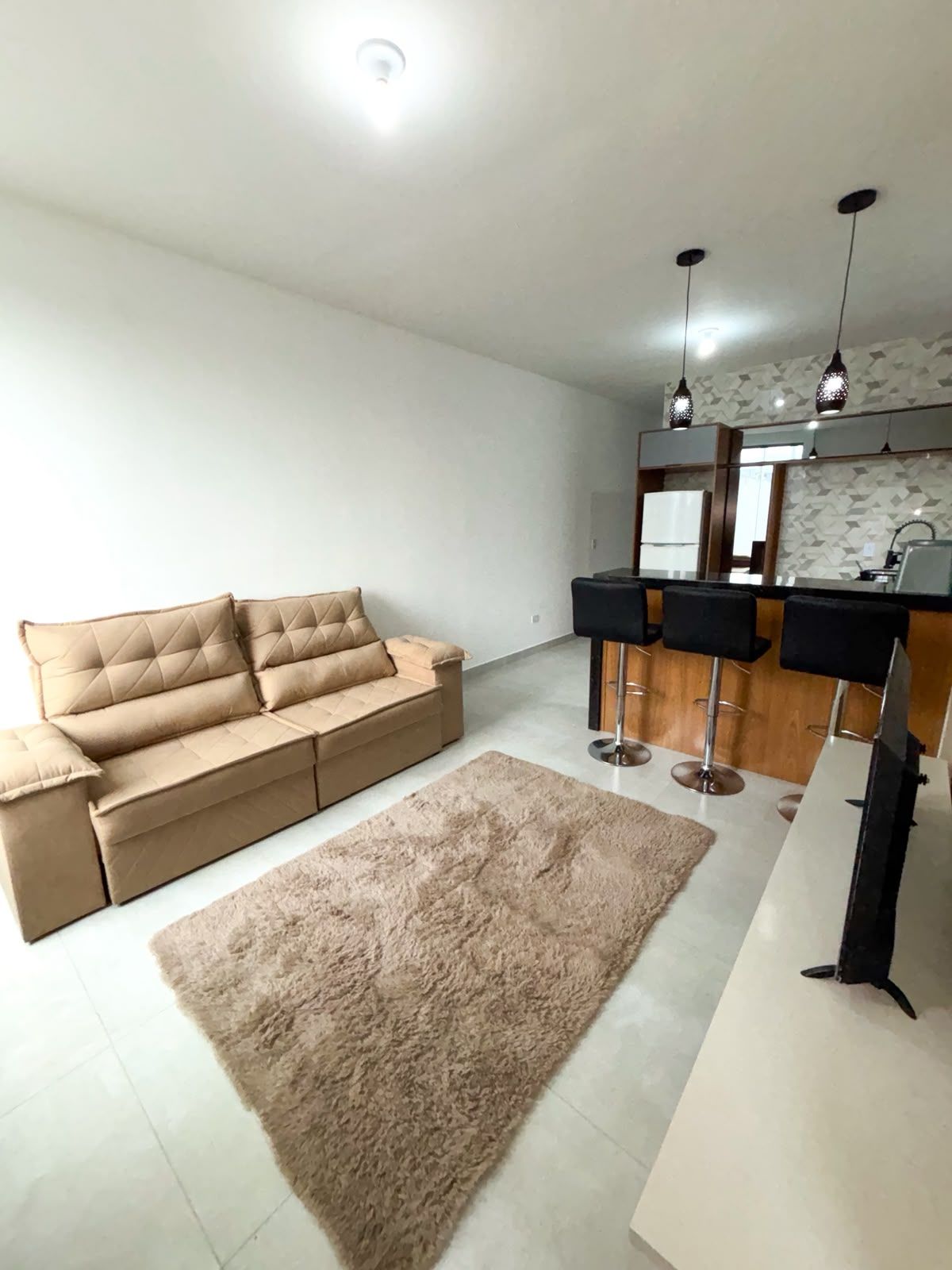 Ein modernes Wohnzimmer mit einem beigefarbenen Sofa, einem flauschigen Teppich und einer Küchenzeile mit Barhocker und Pendelleuchten.