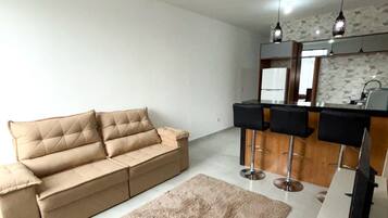 Ein modernes Wohnzimmer mit einem beigefarbenen Sofa, einem flauschigen Teppich und einer Küchenzeile mit Barhocker und Pendelleuchten.