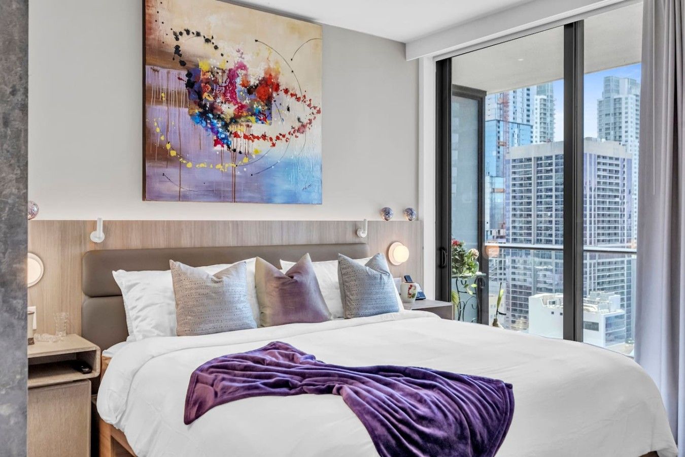 Une chambre d’hôtel moderne avec un grand lit, une vue sur la ville et une peinture abstraite aux couleurs vives accrochée au mur.