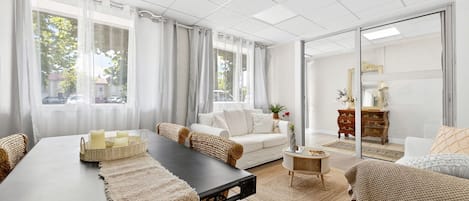 A modern living room with a large table, a sofa, and a coffee table. There is a window with curtains, a mirror, and a dresser with a mirror.