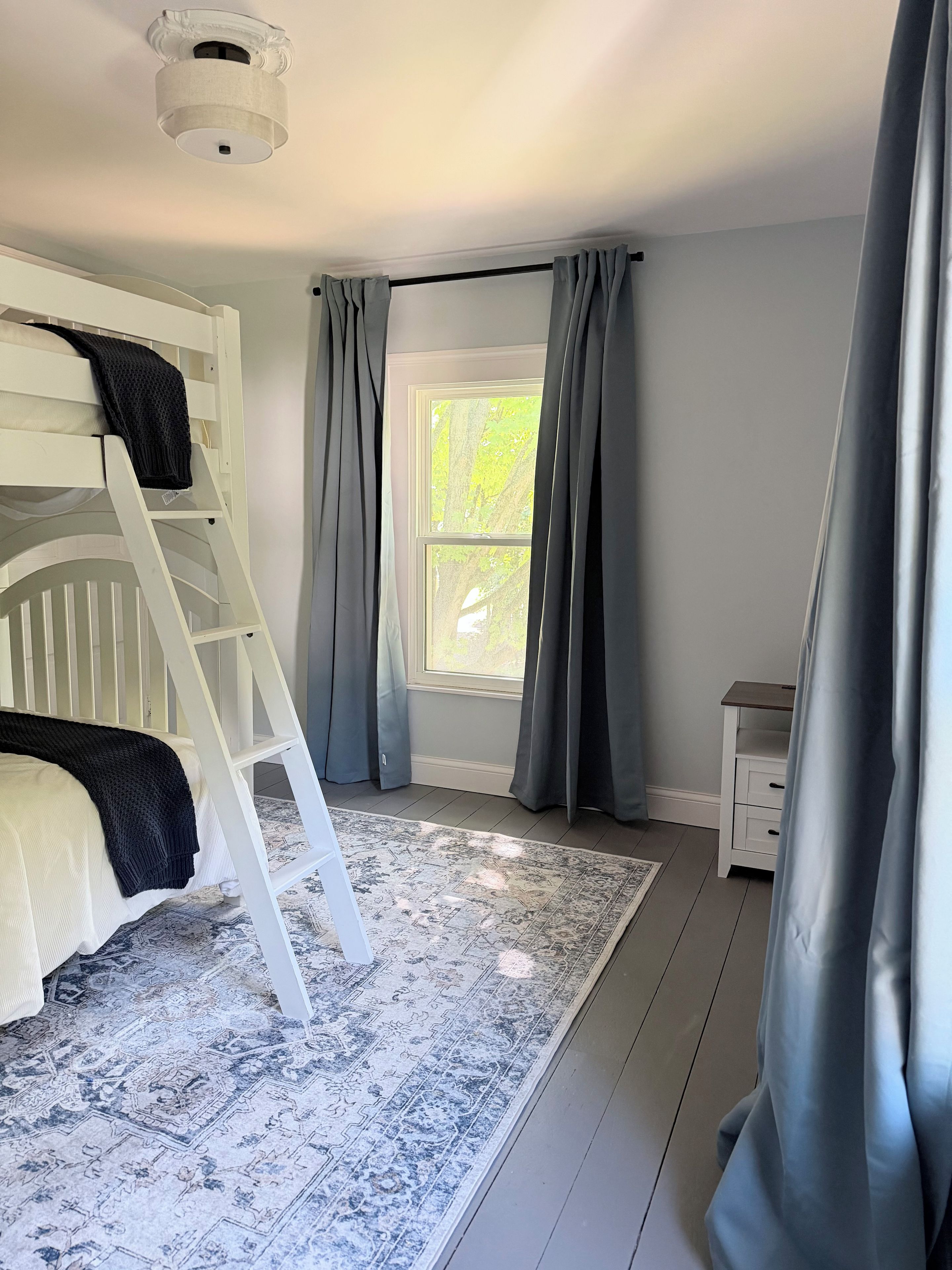A bedroom with a bunk bed, a window with curtains, a nightstand, and a patterned rug.
