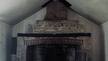 Una habitación con chimenea de piedra, ventilador de techo y cama con manta blanca.