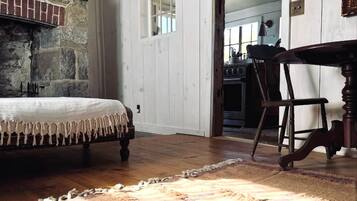 Quarto com uma cama, uma mesa e uma cadeira. Apresenta uma parede de pedra e uma porta de madeira.