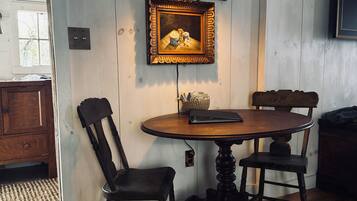 Een eetgedeelte met een ronde houten tafel, twee stoelen, een lijst met een afbeelding en een wandlamp.
