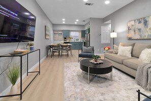 A modern living room with a flat-screen TV, a dining area with a table and chairs, a kitchen with blue cabinets, and a grey sofa with a throw blanket.
