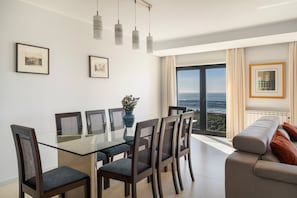 A modern dining area with a glass table and chairs, a view of the ocean through large windows, and a sofa with cushions.