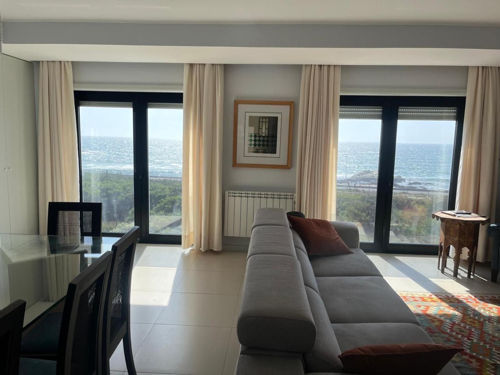 Ein modernes Wohnzimmer mit einer großen Couch, einem Esstisch, Stühlen, einem gerahmten Bild an der Wand und Meerblick durch große Fenster.