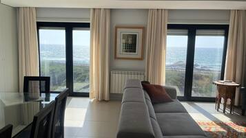 Ein modernes Wohnzimmer mit einer großen Couch, einem Esstisch, Stühlen, einem gerahmten Bild an der Wand und Meerblick durch große Fenster.