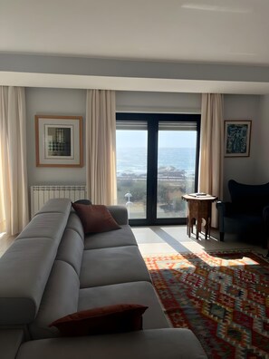 Un salon moderne avec un grand canapé gris, un tapis à motifs et une vue sur la mer par une porte vitrée.