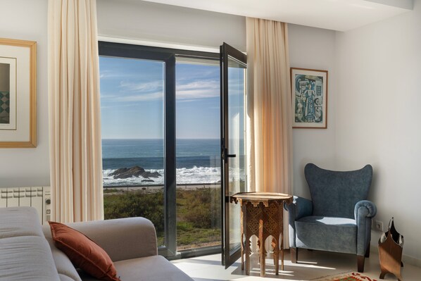 A room with a large sliding glass door leading to a view of the ocean, a blue armchair, a small wooden table, and a framed artwork on the wall.