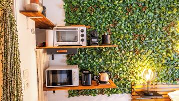 A kitchenette with a microwave, toaster, and kettle on wooden shelves against a wall with a green leafy backdrop.