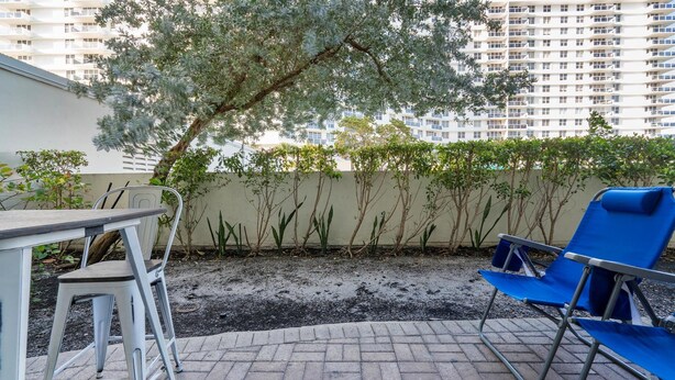 An outdoor patio area with a table and chairs, a blue lounge chair, and a row of small trees against a building wall. - The Tides Hollywood Luxury Suites (Hollywood)