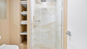 Un bagno con box doccia in vetro, pareti color beige e mensole bianche con asciugamani.