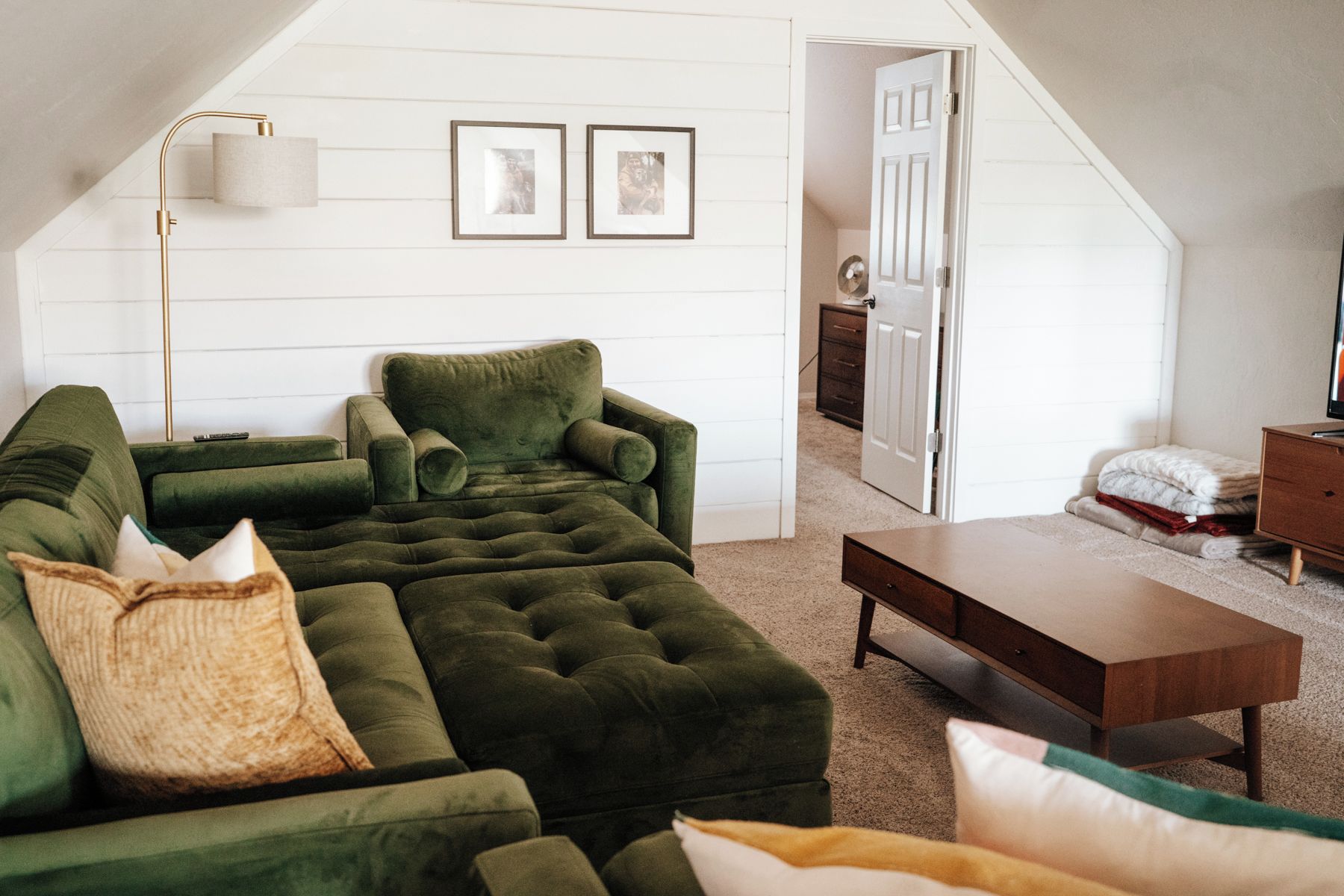 Un salon chaleureux avec un canapé d’angle vert, une table basse en bois et une porte blanche donnant sur une chambre.