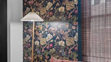 A room with a dark blue floral wallpaper, a brown sofa with cushions, a floor lamp, and a window with blinds.