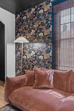 Ein Zimmer mit dunkelblauem Blumenwallpaper, einem braunen Sofa mit Kissen, einer Stehlampe und einem Fenster mit Jalousien.