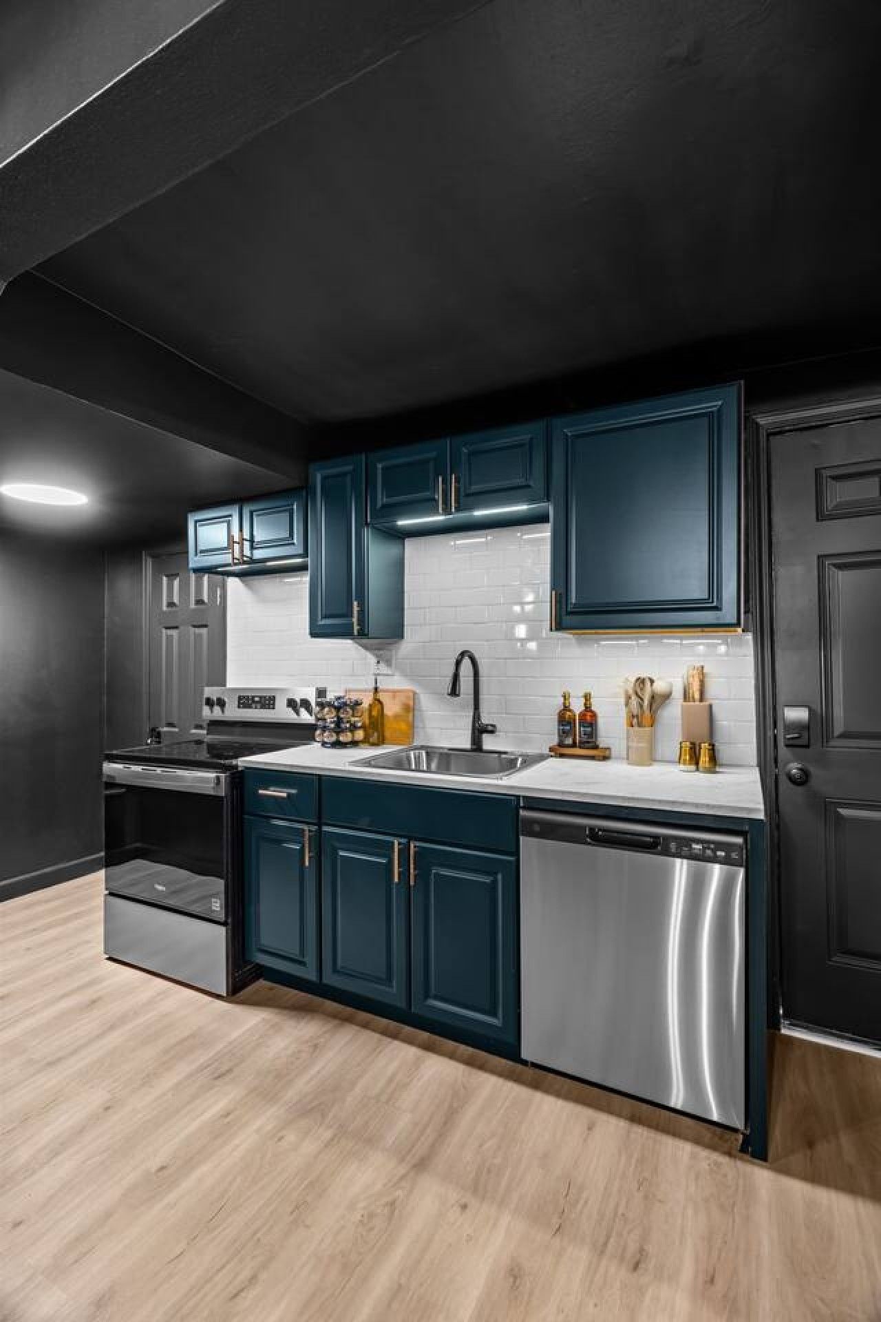 A modern kitchen with dark cabinetry, stainless steel appliances, and a light wood floor.
