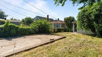 Un jardin à l’arrière d’une maison, avec une terrasse en béton, un petit abri de jardin et la maison en arrière-plan.