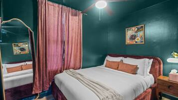 Une chambre à coucher avec un lit, une table de chevet, un miroir, un ventilateur de plafond et un tableau accroché au mur.