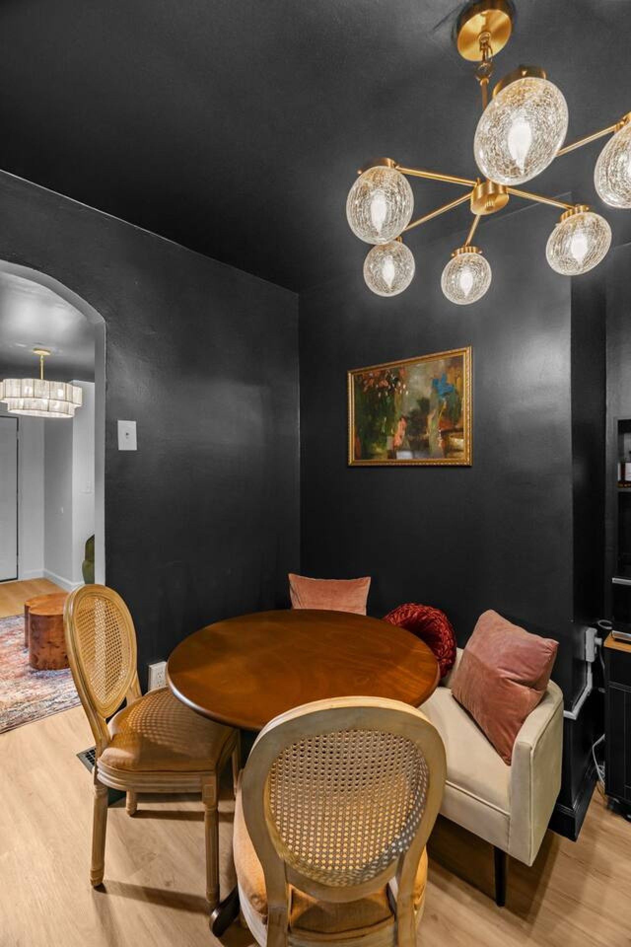 Un coin repas comprenant une table ronde en bois, deux chaises en osier, un canapé avec des coussins et un lustre orné de plusieurs globes de cristal.