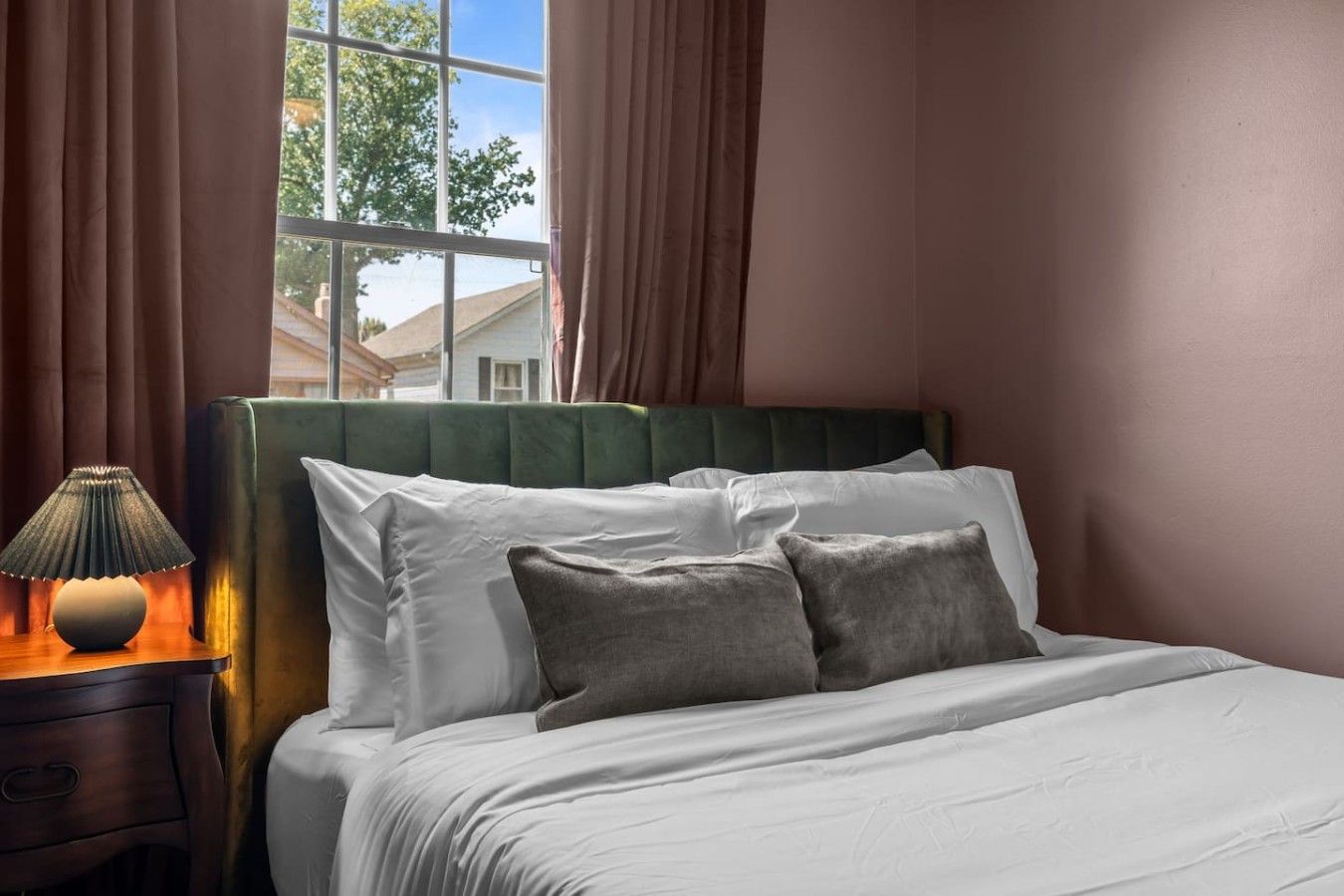 Una cama bien hecha con sábanas blancas y dos almohadas, una lámpara de noche y una ventana con cortinas.