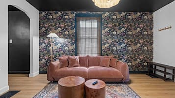 Een kamer met een ruime bank, twee ronde salontafels, een kamerkleed met dessin en bloemenbehang.