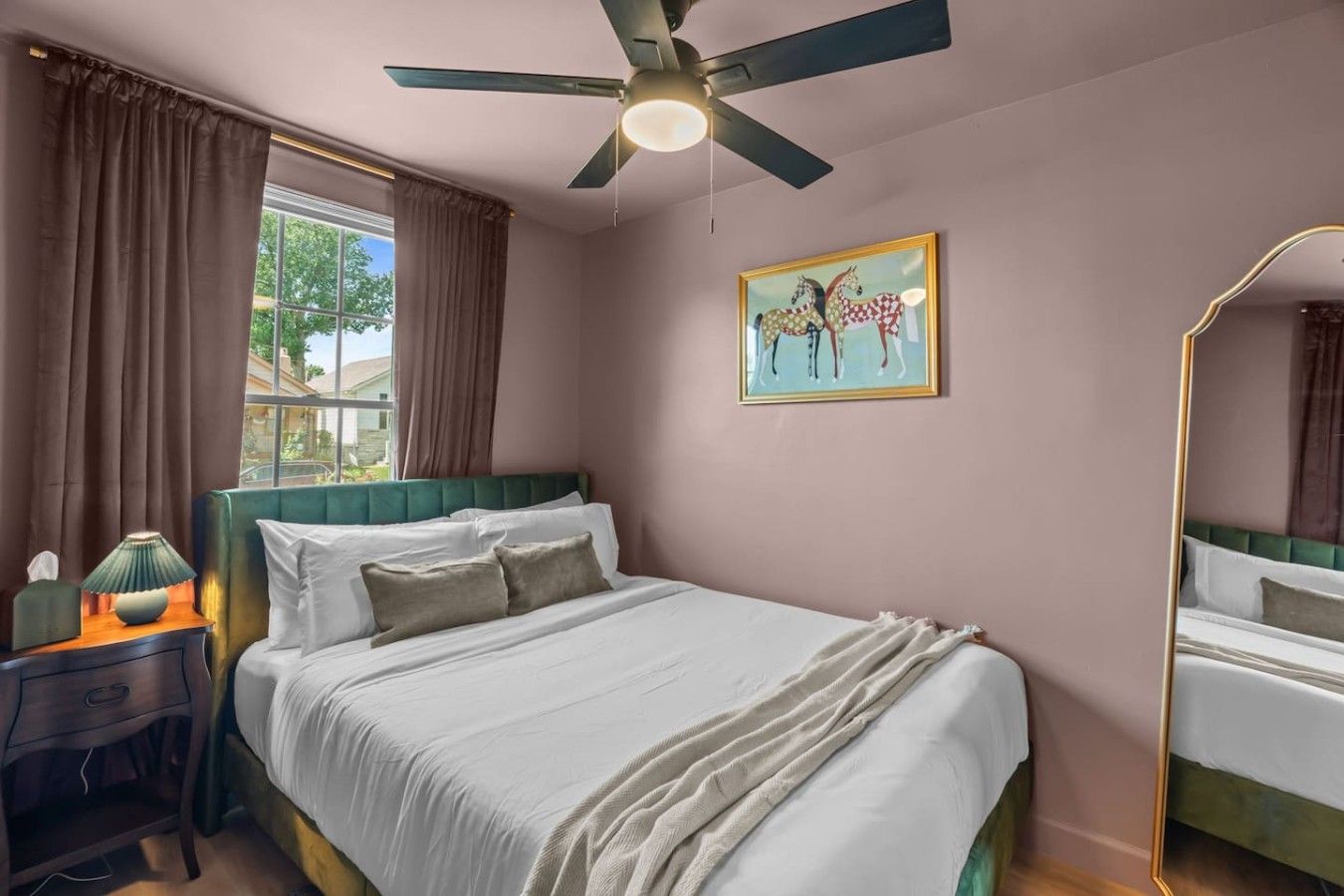Un dormitorio con cama, mesita de noche, lámpara, ventana con cortinas, ventilador de techo y un cuadro en la pared.