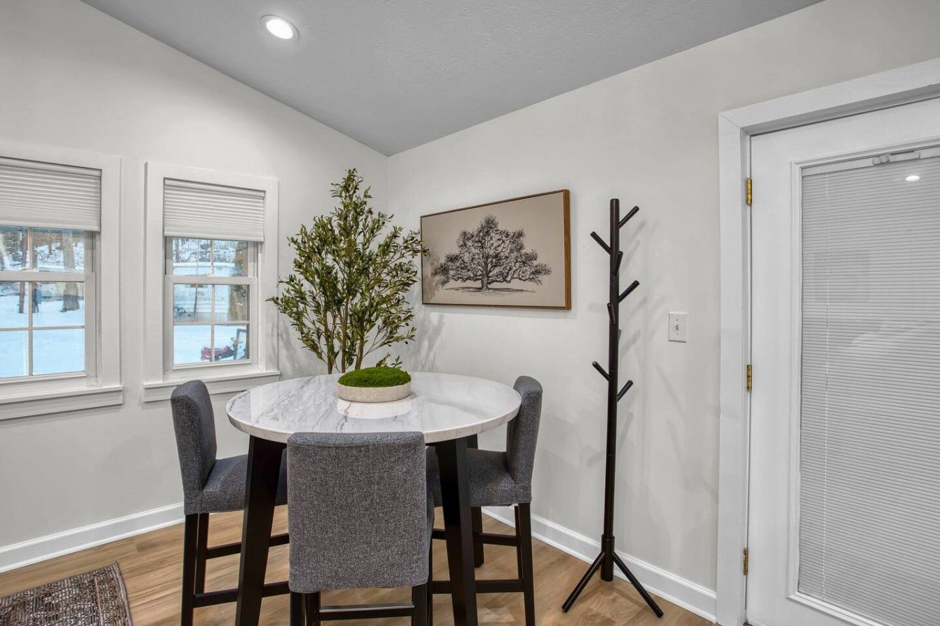 Un coin repas avec une table ronde, quatre chaises, une plante en pot, un porte-manteau et un tableau encadré au mur.