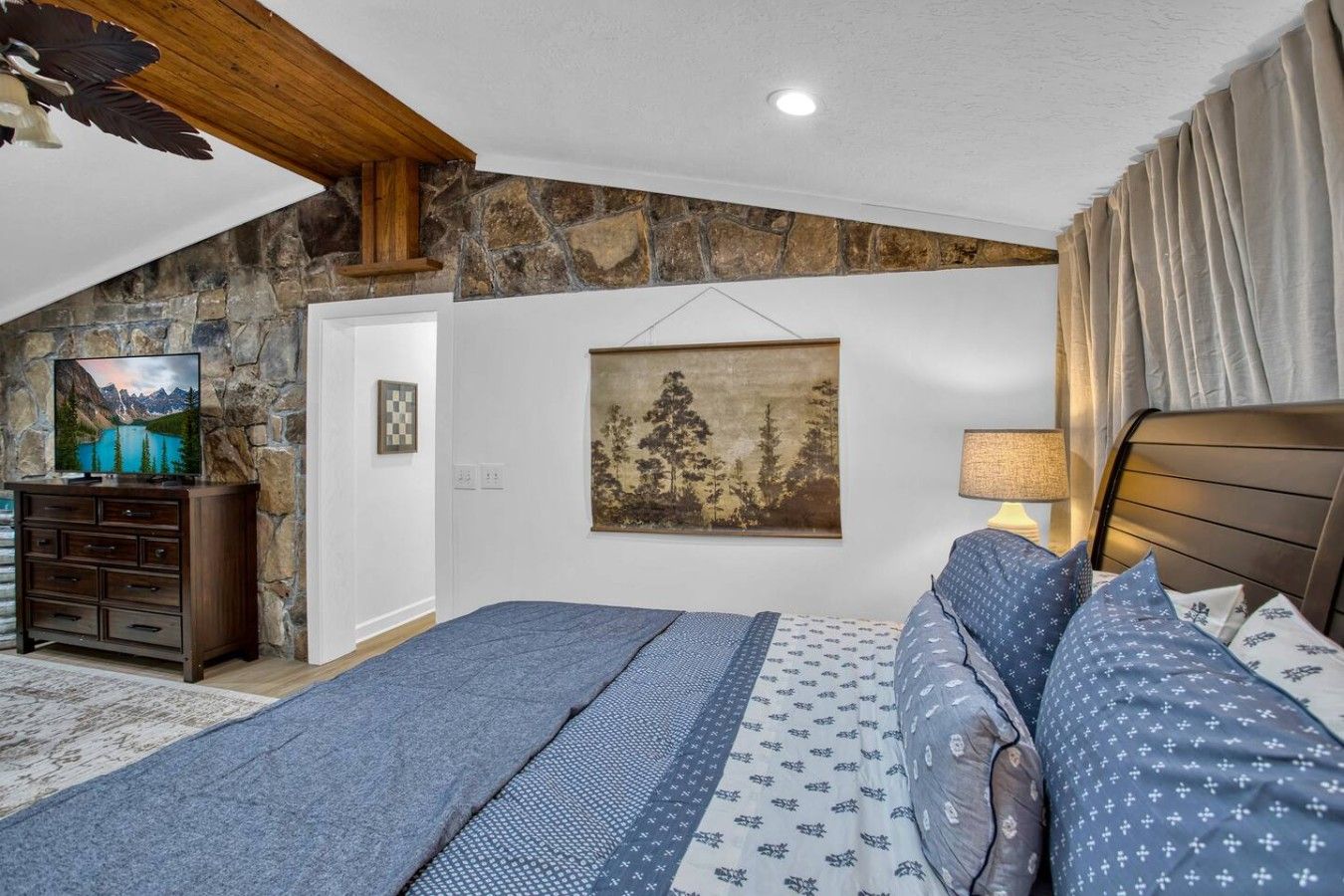 Une chambre avec un mur en pierre, un lit recouvert d’une literie à motifs, un téléviseur posé sur une commode et un tableau accroché au mur.