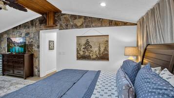 A bedroom with a stone wall, a bed with patterned bedding, a television on a dresser, and a painting on the wall.