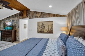 Un dormitorio con pared de piedra, una cama con ropa de cama estampada, un televisor sobre un tocador y un cuadro en la pared.