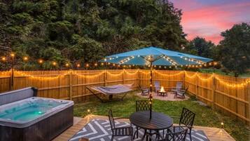A backyard with a hot tub, a deck with a table and chairs, a covered seating area, and string lights.