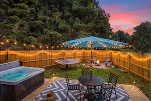 A backyard with a hot tub, a deck with a table and chairs, a covered seating area, and string lights.