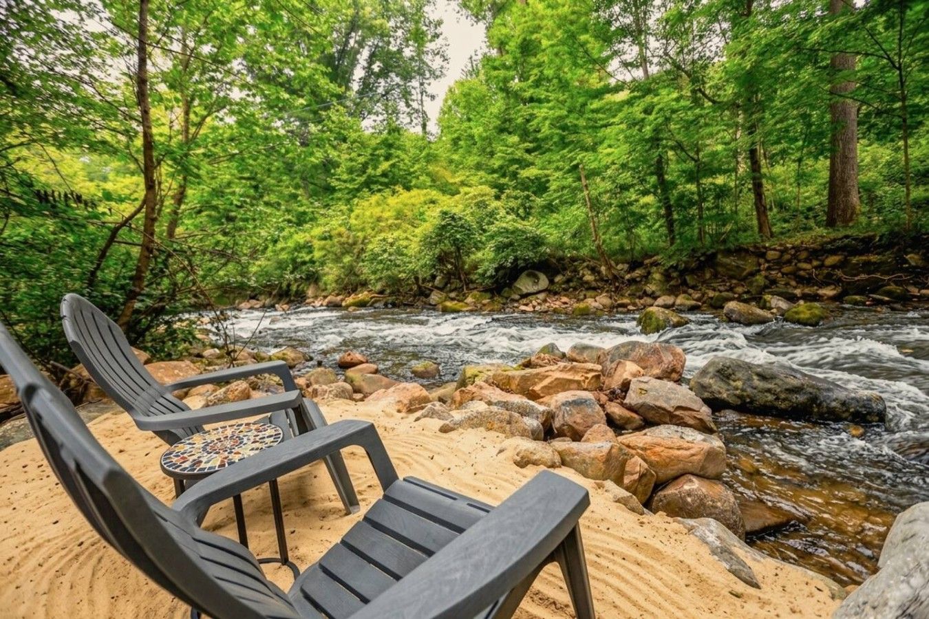 A riverside seating area with chairs and a table, surrounded by rocks and dense greenery.