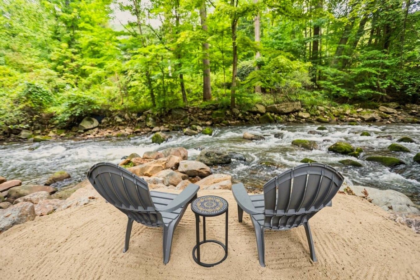 Ein Fluss mit Steinen und Bäumen im Hintergrund, zwei Stühle und ein kleiner Tisch im Vordergrund.