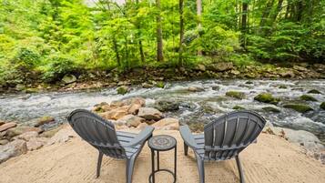 Ein Fluss mit Steinen und Bäumen im Hintergrund, zwei Stühle und ein kleiner Tisch im Vordergrund.