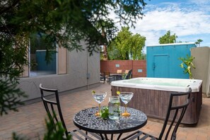 Een terras buiten met een ronde tafel, twee stoelen, een jacuzzi en een blauwe deur op de achtergrond.