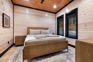 Ein Schlafzimmer mit Bett, Nachttisch, Kommode und großen Fenstern.